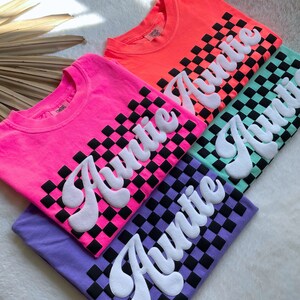 May include: Four t-shirts with a checkered pattern and the word "Auntie" in a retro font. The shirts are in pink, orange, green, and purple.