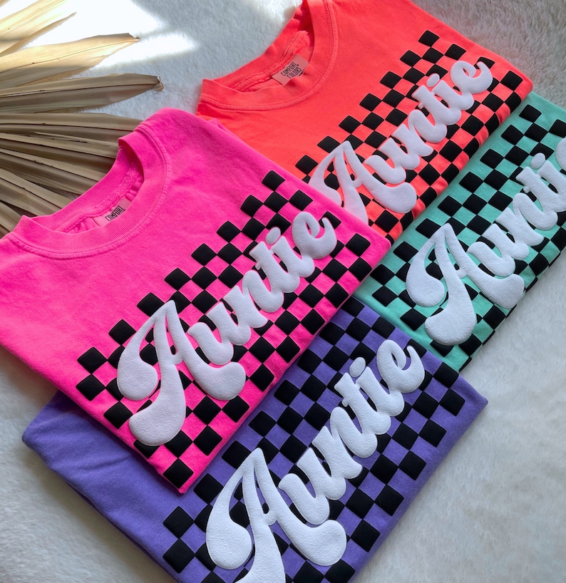 May include: Four t-shirts with a checkered pattern and the word "Auntie" in a retro font. The shirts are in pink, orange, green, and purple.