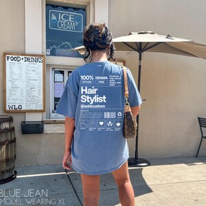 May include: A blue t-shirt with a white graphic that says "100% Drama Styled Free Hair Stylist @addcustom". The graphic is a label with a barcode and icons.