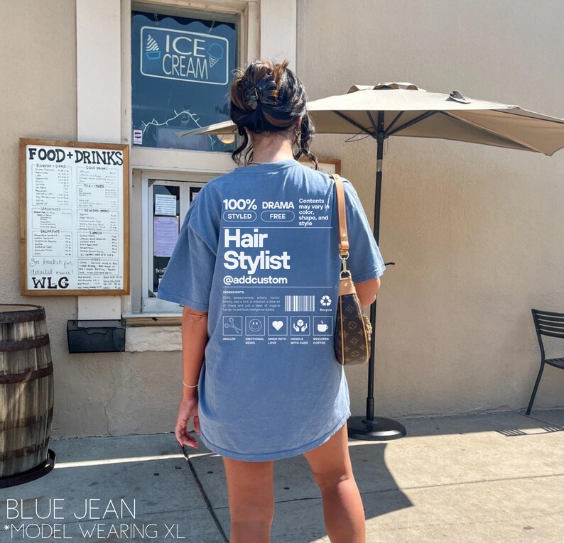 May include: A blue t-shirt with a white graphic that says "100% Drama Styled Free Hair Stylist @addcustom". The graphic is a label with a barcode and icons.