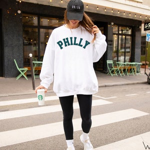 May include: A white sweatshirt with the word "PHILLY" in green lettering on the front. The person is wearing black leggings and a black baseball cap with a white logo.