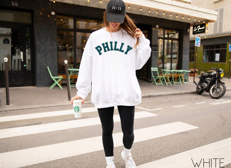 May include: A white sweatshirt with the word "PHILLY" in green lettering on the front. The person is wearing black leggings and a black baseball cap with a white logo.