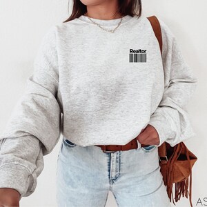 May include: A gray sweatshirt with the word "Realtor" and a barcode printed on the front. The sweatshirt is worn by a person wearing blue jeans and a brown belt.