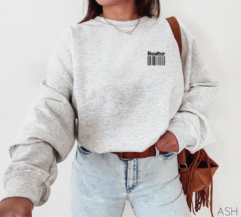 May include: A gray sweatshirt with the word "Realtor" and a barcode printed on the front. The sweatshirt is worn by a person wearing blue jeans and a brown belt.