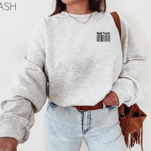 May include: A light gray sweatshirt with the text "Nail Tech" above a barcode design. The sweatshirt has long sleeves and a crew neck. The word "ASH" is in the upper left corner.