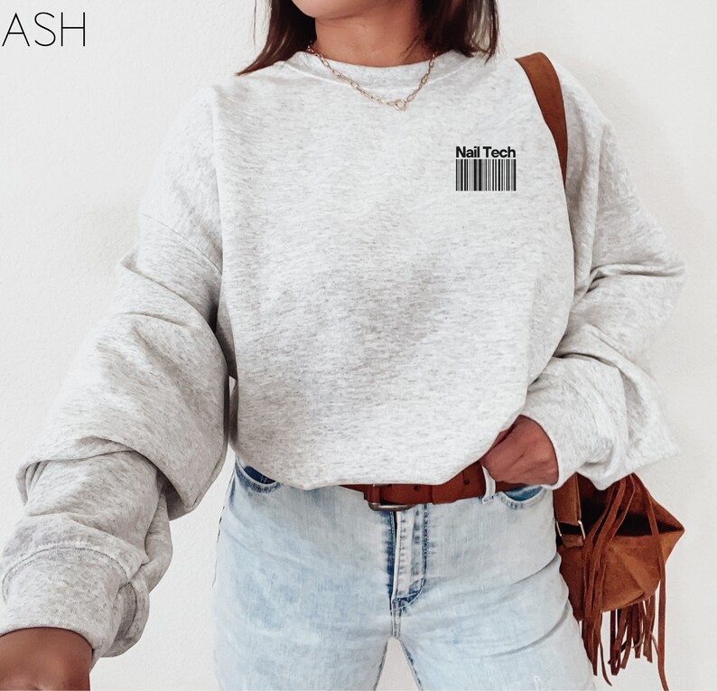 May include: A light gray sweatshirt with the text "Nail Tech" above a barcode design. The sweatshirt has long sleeves and a crew neck. The word "ASH" is in the upper left corner.
