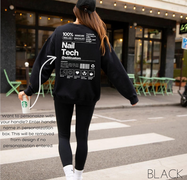May include: Black crewneck sweatshirt with white text that reads "Nail Tech @addcustom" and a graphic on the back. The sweatshirt also has additional text and icons. The person is wearing black leggings and a black cap.