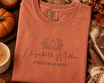 May include: A terracotta-colored short-sleeve t-shirt featuring a black line-art graphic of a camera surrounded by flowers. Below the graphic, the text reads Elizabeth Miller PHOTOGRAPHY in a combination of script and block lettering.