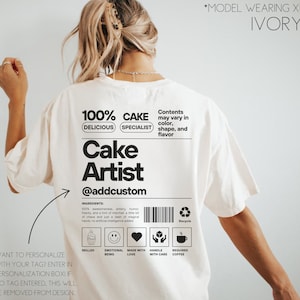May include: White t-shirt with a black graphic design that says "100% Delicious Cake Specialist" and "Cake Artist @addcustom". The graphic also includes a barcode, a recycle symbol, and icons for skilled, emotional being, made with love, handle with care, and requires coffee.