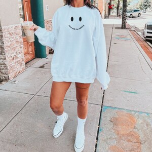May include: White sweatshirt with a smiley face design and the text "can't talk I'm editing" in black. The sweatshirt is worn with white high-top sneakers and white socks.