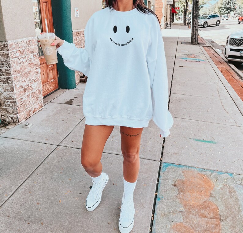 May include: White sweatshirt with a smiley face design and the text "can't talk I'm editing" in black. The sweatshirt is worn with white high-top sneakers and white socks.