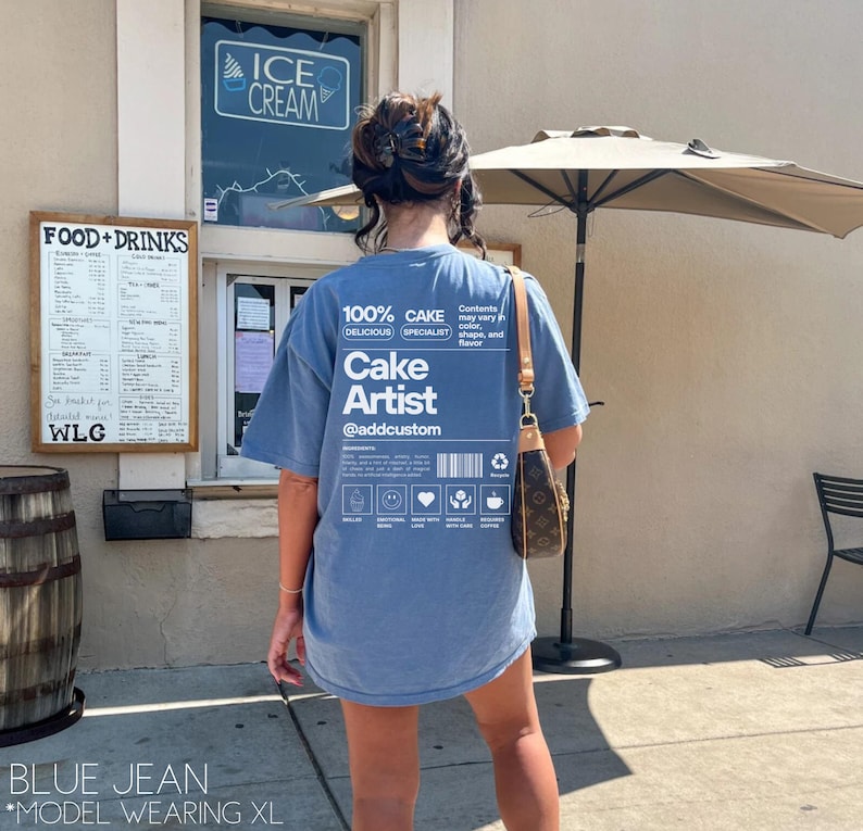 May include: A blue jean t-shirt with a white graphic design that says "100% CAKE DELICIOUS SPECIALIST Cake Artist @addcustom". The graphic design looks like a product label with a barcode and a list of ingredients.