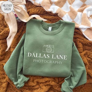 Può includere: Felpa verde militare con la scritta "DALLAS LANE PHOTOGRAPHY" e una grafica di una fotocamera. La felpa girocollo è esposta su una superficie marrone strutturata.