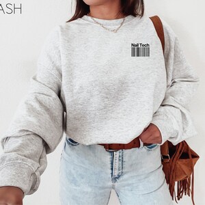 May include: A light gray sweatshirt with the text "Nail Tech" above a barcode design. The sweatshirt has long sleeves and a crew neck. The model is wearing light wash jeans and a brown belt.