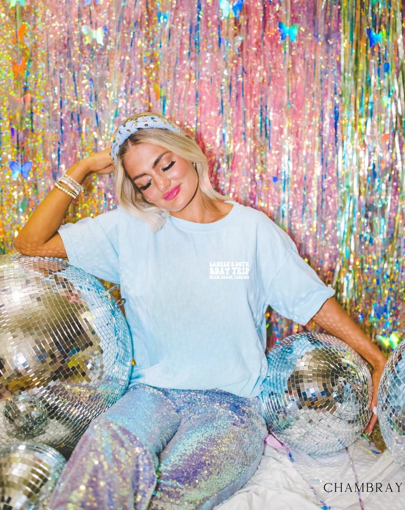 May include: A woman wearing a light blue t-shirt with the text "Lauren's South Bay Trip" and a sparkly iridescent skirt. She is sitting in front of a pink and blue iridescent background with disco balls.