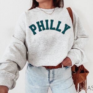 May include: A gray sweatshirt with the word "PHILLY" printed in teal blue letters on the front. The sweatshirt has a relaxed fit and is worn by a person with a brown leather bag.