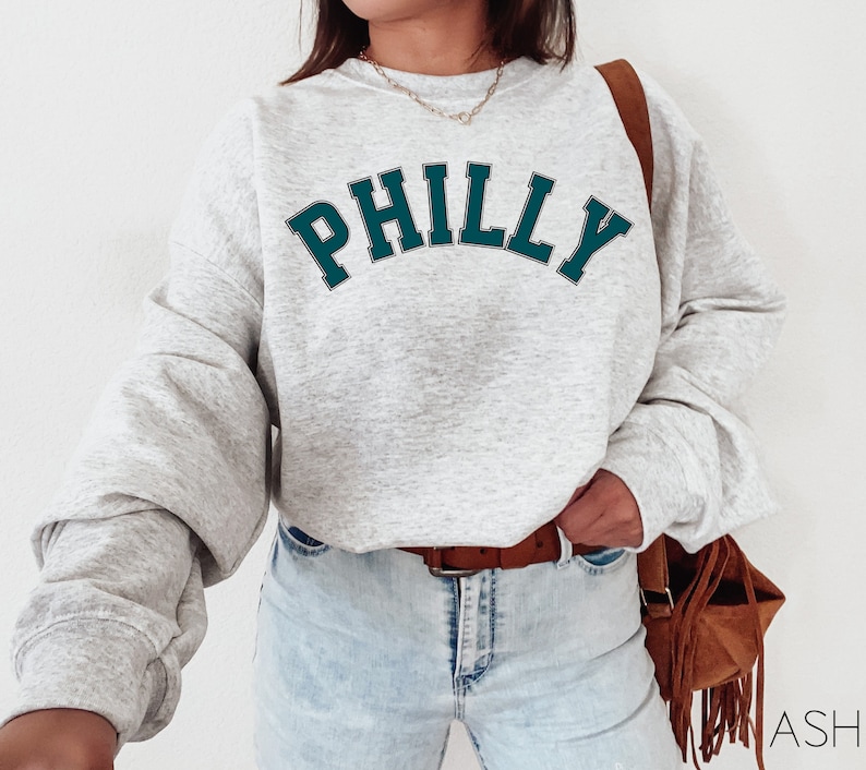 May include: A gray sweatshirt with the word "PHILLY" printed in teal blue letters on the front. The sweatshirt has a relaxed fit and is worn by a person with a brown leather bag.