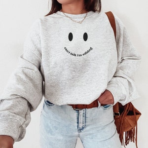May include: A light gray sweatshirt with a smiley face design and the text "can't talk I'm editing" in black. The sweatshirt has long sleeves and a crew neck. The model is wearing light blue jeans and a brown belt.