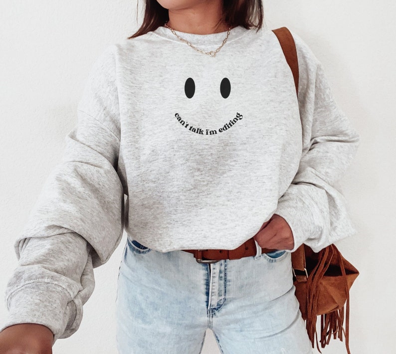 May include: A light gray sweatshirt with a smiley face design and the text "can't talk I'm editing" in black. The sweatshirt has long sleeves and a crew neck. The model is wearing light blue jeans and a brown belt.