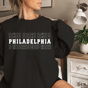 Puede incluir: Sudadera negra con la palabra "PHILADELPHIA" en letras blancas. La palabra se repite varias veces, creando un patr&oacute;n. La sudadera es de cuello redondo.