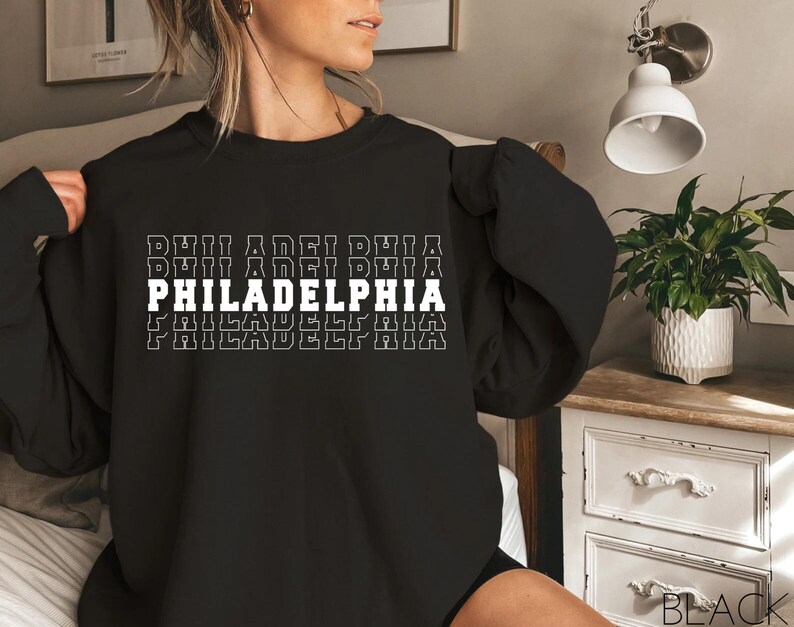 Puede incluir: Sudadera negra con la palabra "PHILADELPHIA" en letras blancas. La palabra se repite varias veces, creando un patr&oacute;n. La sudadera es de cuello redondo.