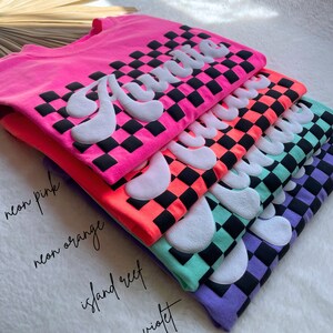 May include: A stack of five t-shirts with the word "Auntie" printed in white on a black and white checkered background. The shirts are in different colors: neon pink, neon orange, island reef, and violet.
