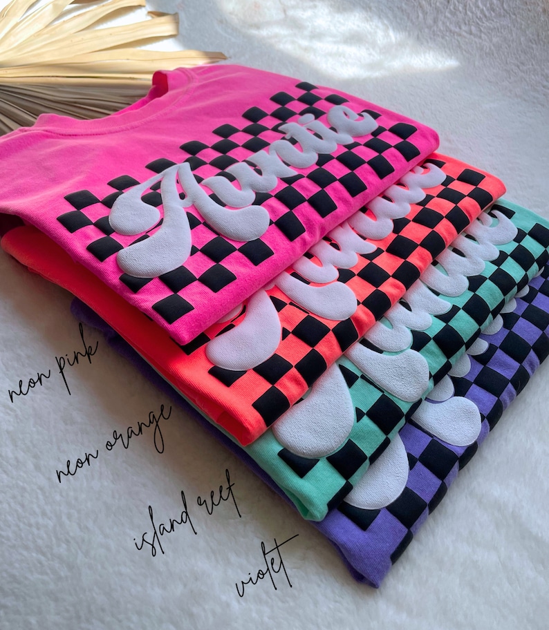 May include: A stack of five t-shirts with the word "Auntie" printed in white on a black and white checkered background. The shirts are in different colors: neon pink, neon orange, island reef, and violet.