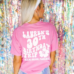 May include: Pink t-shirt with white text that says "Lauren's 30th Birthday Trip Miami Beach, Florida" with a white hibiscus flower graphic.