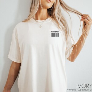 May include: Ivory t-shirt with the text "Hairstylist" and a barcode printed on the front. The model is wearing a size XL.