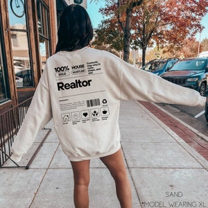 May include: A sand-colored sweatshirt with a black graphic design that reads "100% House Hustler Sold Realtor". The graphic also includes a barcode and a list of ingredients, including "requires coffee".