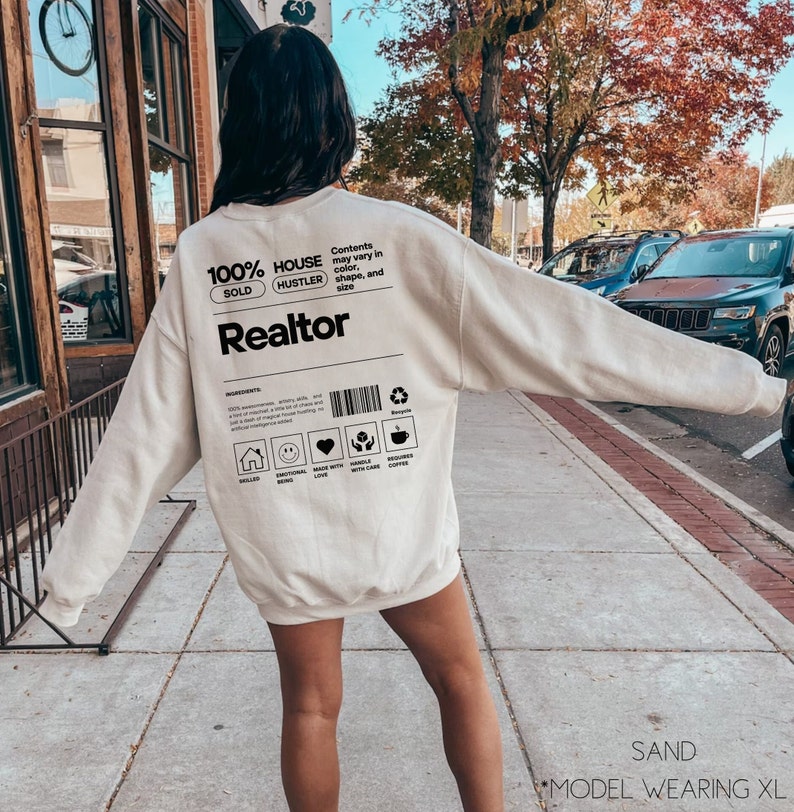 May include: A sand-colored sweatshirt with a black graphic design that reads "100% House Hustler Sold Realtor". The graphic also includes a barcode and a list of ingredients, including "requires coffee".