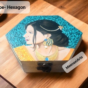 May include: A hexagonal wooden box featuring a painted image of a woman in a yellow and blue sari, adorned with jewellery. The box has a hinged lid and a metal clasp. The text "MeenaxiArts" is printed on the bottom right corner of the box.