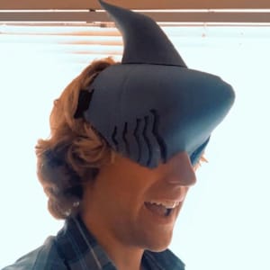 May include: A blue shark-shaped headpiece with a dorsal fin and gill slits. The headpiece is worn by a person, creating a playful and whimsical look. The person is wearing a plaid shirt.