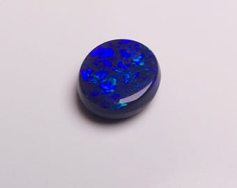 May include: A polished, oval-shaped opal gemstone. The opal has a dark blue base color with vibrant flashes of blue and green throughout. The stone is set against a plain white background, highlighting its natural beauty.
