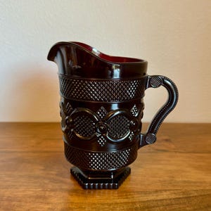 May include: A dark red glass pitcher with a decorative pattern. The pitcher has a handle and a spout for pouring. The design includes diamond and oval shapes. The pitcher is sitting on a wooden surface.