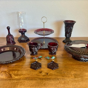 May include: A collection of vintage ruby red glass serving pieces, including a bell, a two-tiered server, a vase, a pitcher, a cup, a platter, a cake stand with a knife, and two ornaments. The pieces are arranged on a wooden surface.