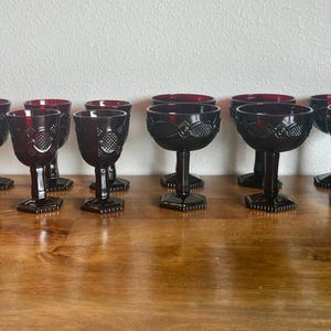 May include: A collection of dark red goblets with intricate embossed designs. The stemmed glasses have a deep red interior and sit on a wooden surface. The goblets are arranged in rows, showcasing their decorative details.