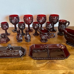 May include: A set of red glass tableware, including a gravy boat, a butter dish, a serving dish, and various cups and glasses. The tableware is decorated with a raised pattern of leaves and flowers.