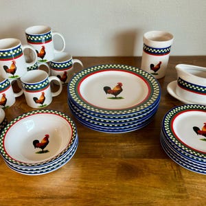 May include: A collection of white ceramic tableware with a rooster motif. Includes plates, bowls, mugs, a gravy boat, and a sugar bowl. Each piece features a red, green, and blue checkered border and a rooster illustration.