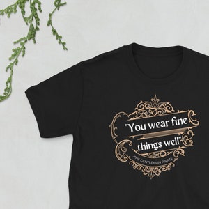 以下が含まれることがあります： 「You wear fine things well」と「The Gentleman Pirate」の文字が入った、金色のグラフィックデザインが施された黒いTシャツです。