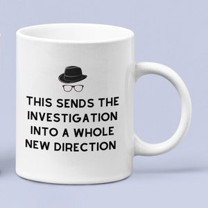 Puede incluir: Dos tazas de cerámica blanca con asas negras. Cada taza presenta una ilustración en blanco y negro de una persona con un fedora y gafas. El texto en cada taza dice "This sends the investigation into a whole new direction."