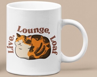 Könnte beinhalten: Zwei weiße Keramikbecher mit einer Illustration einer schwarzen und orangefarbenen Katze. Der Text auf den Bechern lautet "Live, Lounge, Loaf".