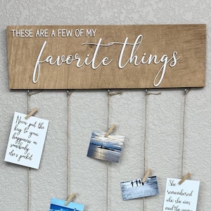 Può includere: Un cartello di legno con il testo "These are a few of my favorite things" in lettere bianche. Il cartello ha corde tese attraverso di esso con mollette che tengono foto e citazioni ispiratrici.