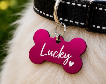 Puede incluir: Una etiqueta para mascotas en forma de hueso magenta con la palabra "Lucky" en escritura blanca y un pequeño corazón blanco. La etiqueta cuelga de un collar negro para perros con costuras blancas.