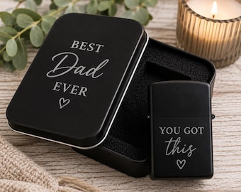 May include: A black metal gift set with a lighter and a tin. The tin lid reads "BEST Dad EVER" with a heart symbol. The lighter says "YOU GOT this" with a heart. The set is on a wooden surface.
