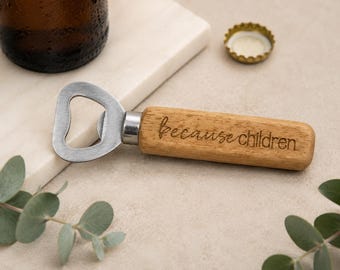 May include: A wooden bottle opener with a stainless steel head. The handle is engraved with the words "Because children". A brown bottle and a bottle cap are in the background. The opener is on a light-colored surface.