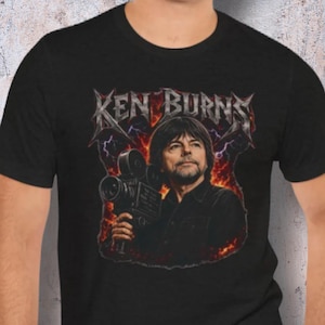 May include: Black t-shirt featuring a portrait of Ken Burns holding a vintage film camera. The design includes the name "KEN BURNS" in a stylized font with lightning and flames, creating a vintage, artistic aesthetic.