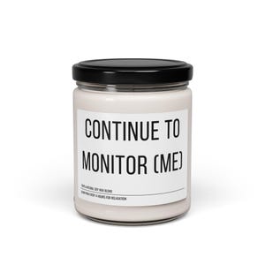 May include: A white candle in a glass jar with a black lid. The label reads "CONTINUE TO MONITOR (ME)". The bottom of the label says "100% NATURAL SOY WAX BLEND" and "BURN FOR EVERY 4 HOURS FOR RELAXATION".
