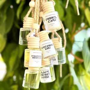 May include: A collection of small, clear glass car air fresheners hanging from a beige rope with wooden beads. Each bottle has a wooden cap and a label with a different scent name, including "Funnel Cake" and "Black Cherry Bomb."