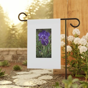 Op de afbeelding: Een witte tuinvaandel met een afbeelding van een paarse irisbloem met witte strepen. De vlag wordt gepresenteerd op een zwarte metalen standaard in een tuin, omringd door groen en stenen paden. De iris staat in volle bloei.